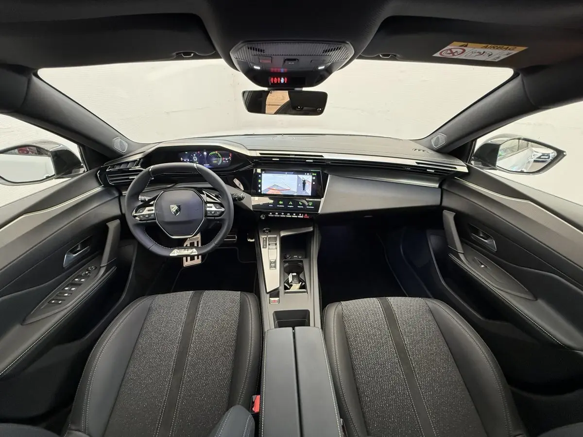 Vue intérieure avant de la Peugeot 308 Hybrid 136 e-DCS6 GT 2024, tableau de bord moderne avec écran tactile central et volant cuir noir.