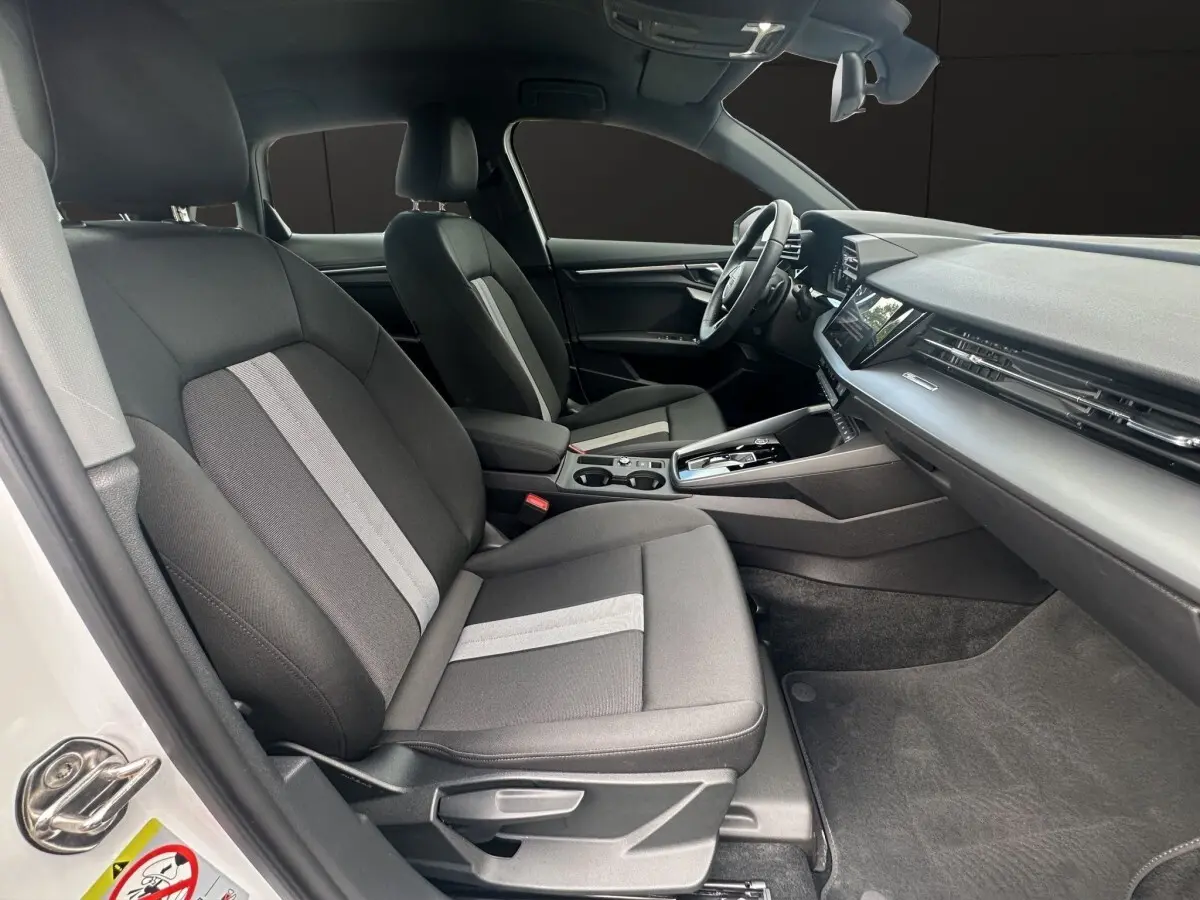 Vue intérieure côté gauche de l’Audi A3 2024 blanche, montrant les sièges tissu gris avec bande centrale claire et tableau de bord moderne.