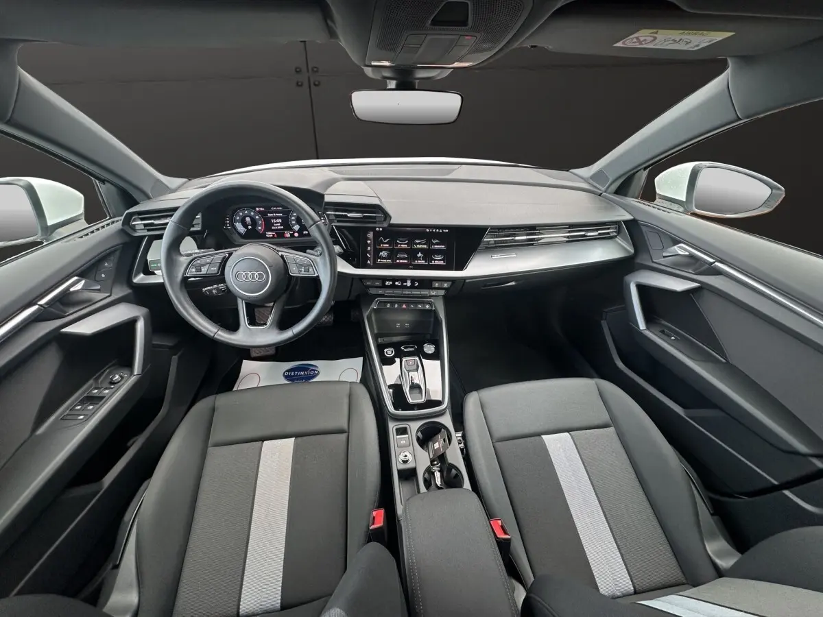Vue intérieure avant de l'Audi A3 2024 blanche, avec tableau de bord numérique et sièges noirs à bande grise centrale.