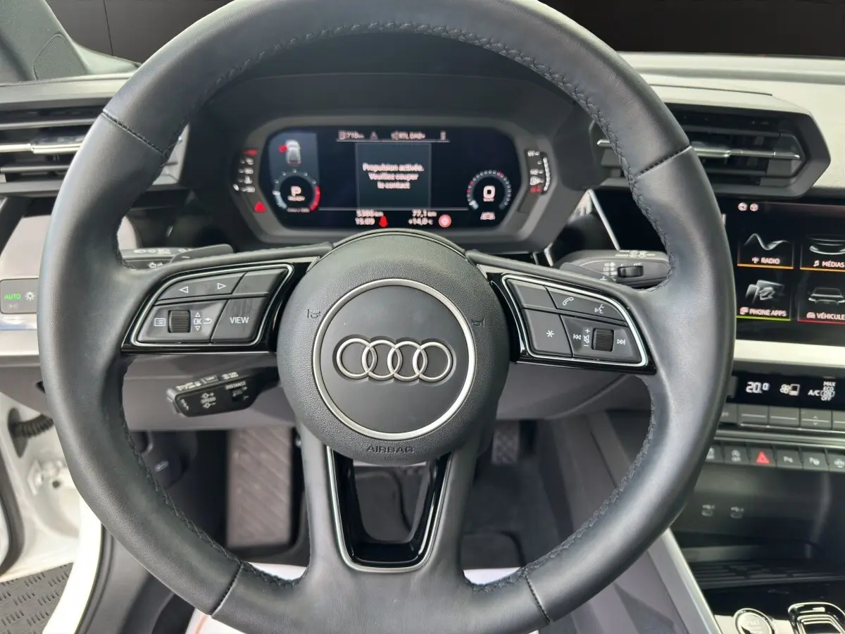 Vue centrée sur le volant noir de l'Audi A3 2024 blanc, avec tableau de bord numérique et commandes intégrées visibles.