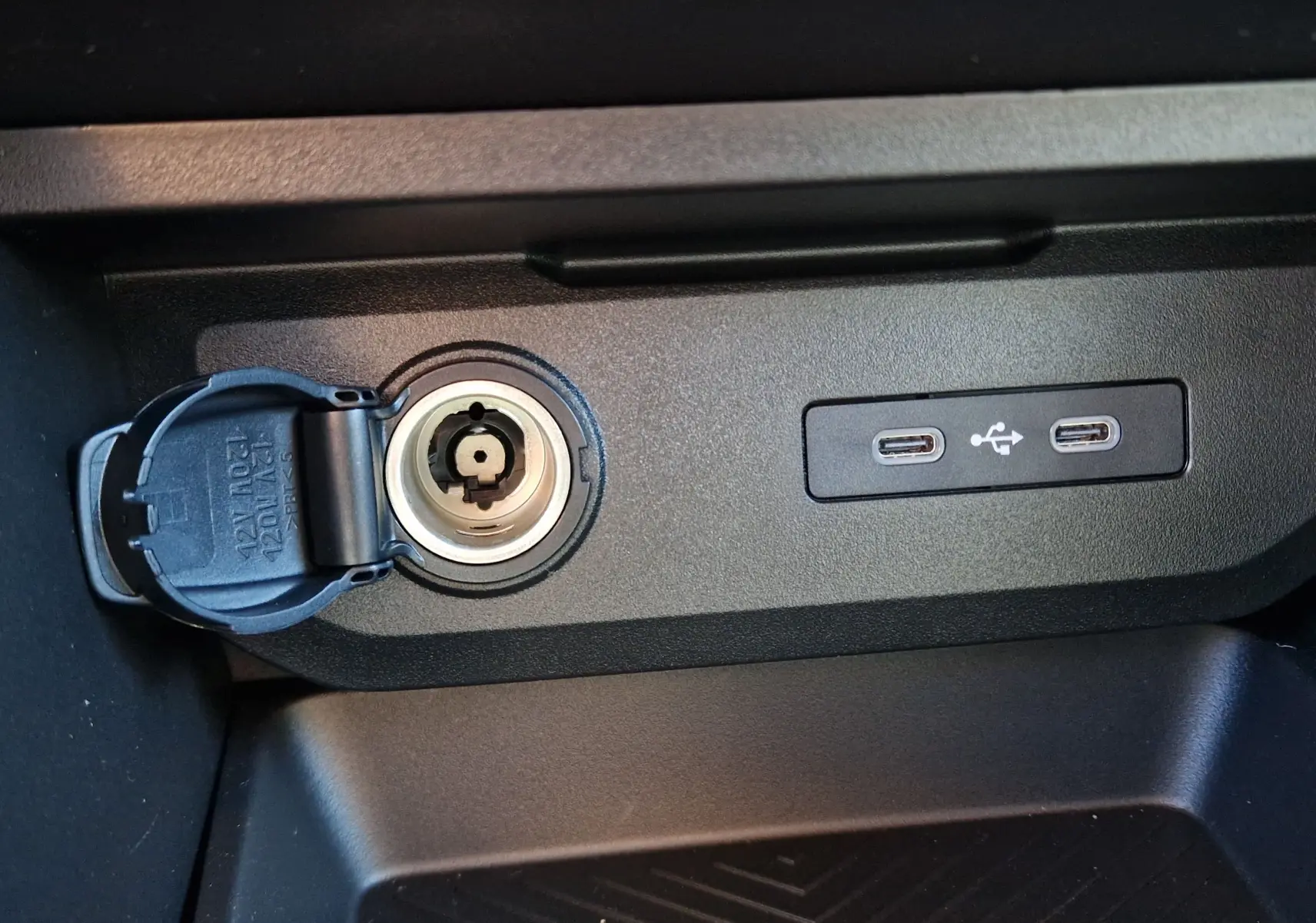 Gros plan intérieur sur la prise 12V ouverte et deux ports USB-C du Dacia Duster 1.8 Hybrid 155 Extreme, finition noire.