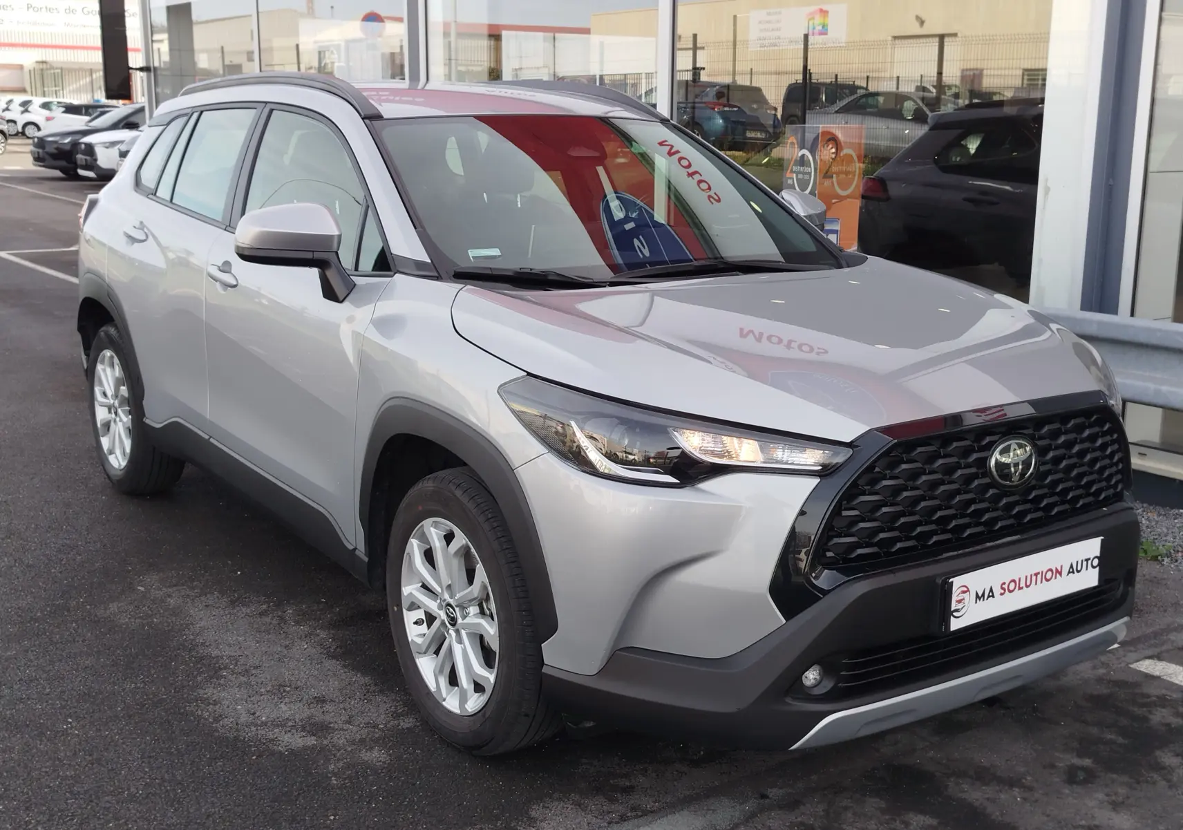 Toyota Corolla Cross gris acier vue 3/4 avant droit, avec calandre noire et phares allumés, stationnée devant un garage.