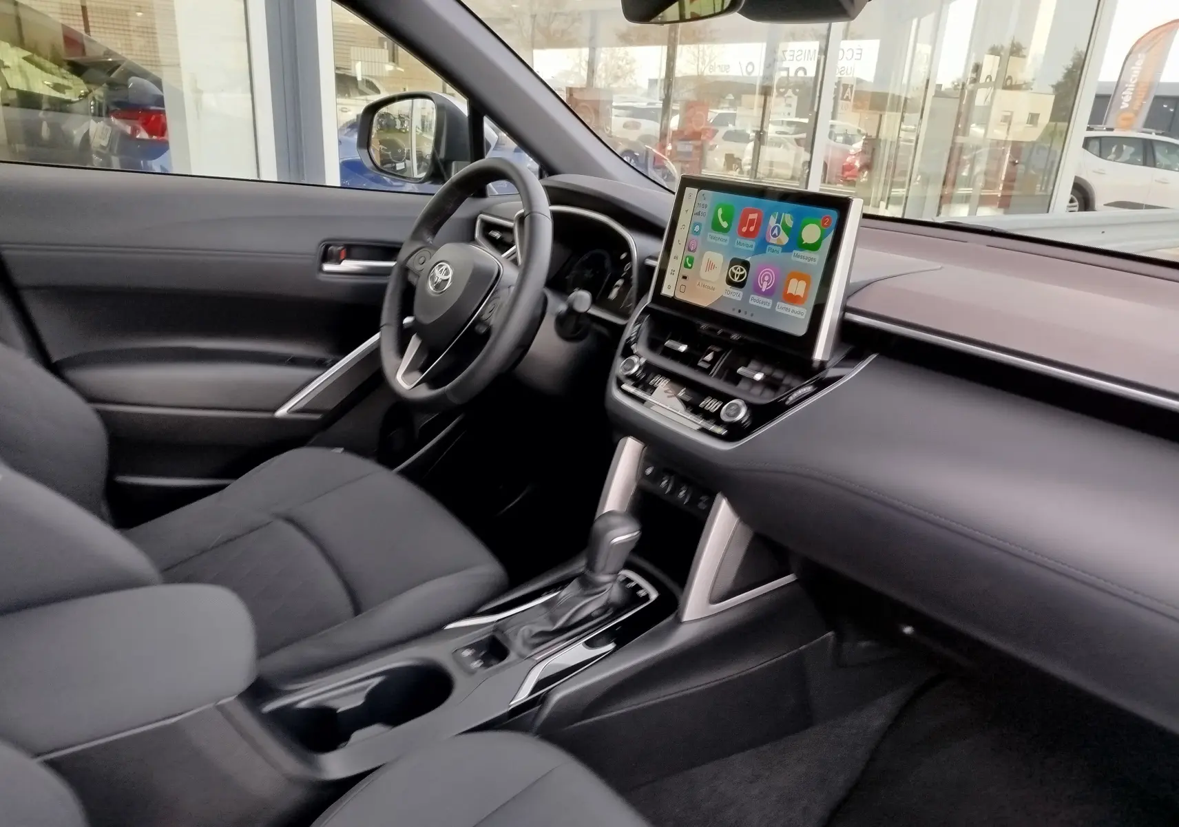 Intérieur avant du Toyota Corolla Cross 2025, tableau de bord noir avec écran tactile central et sièges tissu gris foncé.