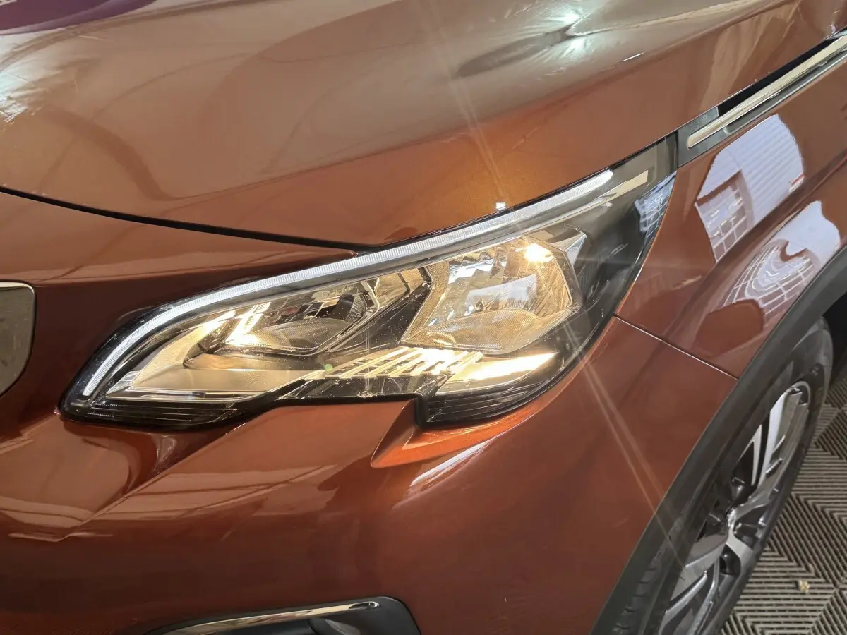 Gros plan sur le phare avant droit allumé d’un Peugeot 3008 orange métallisé, avec partie de la roue visible.