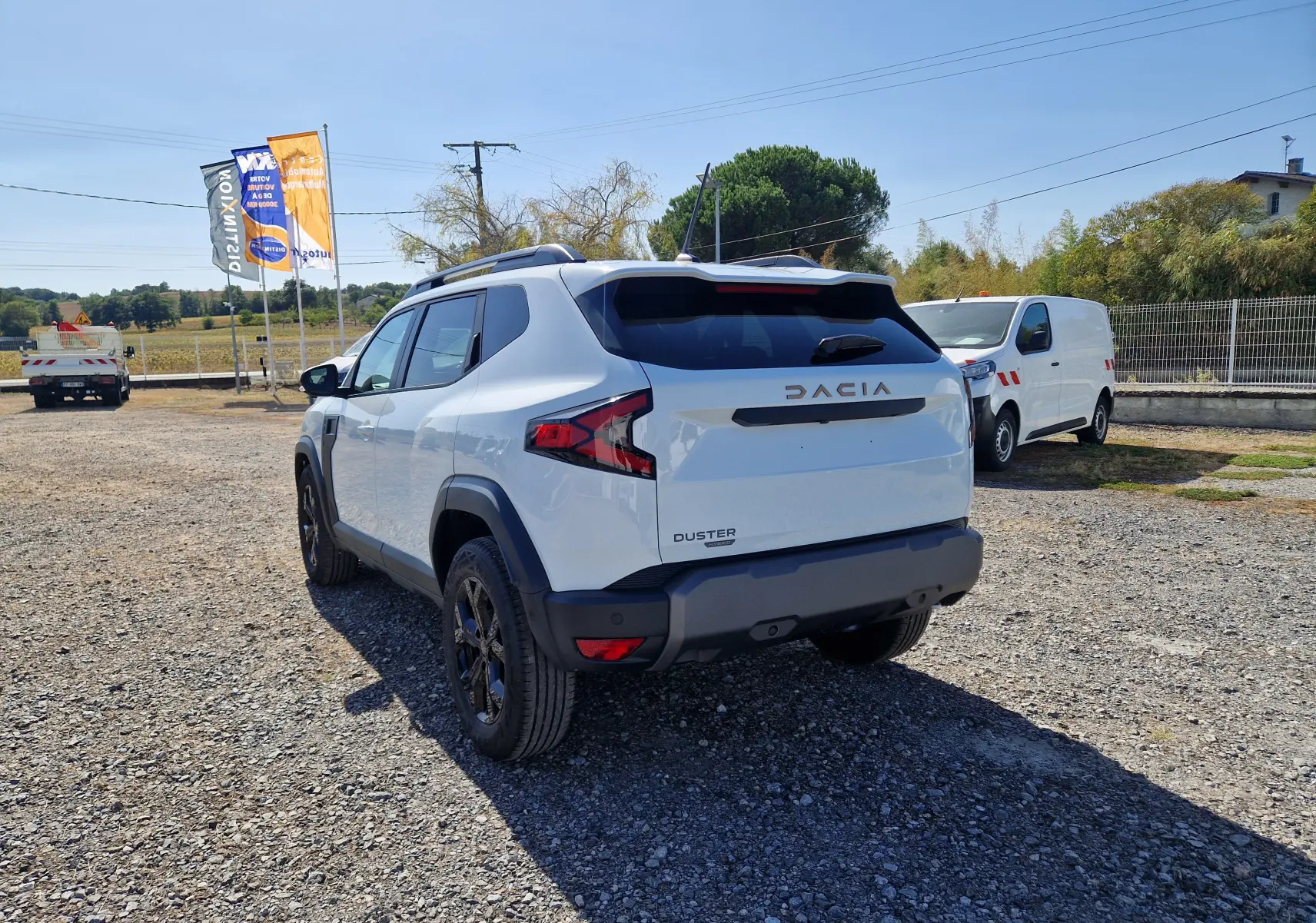 Vue 3/4 arrière droite du Dacia Duster blanc 2026 hybride, avec jantes noires et barres de toit visibles.
