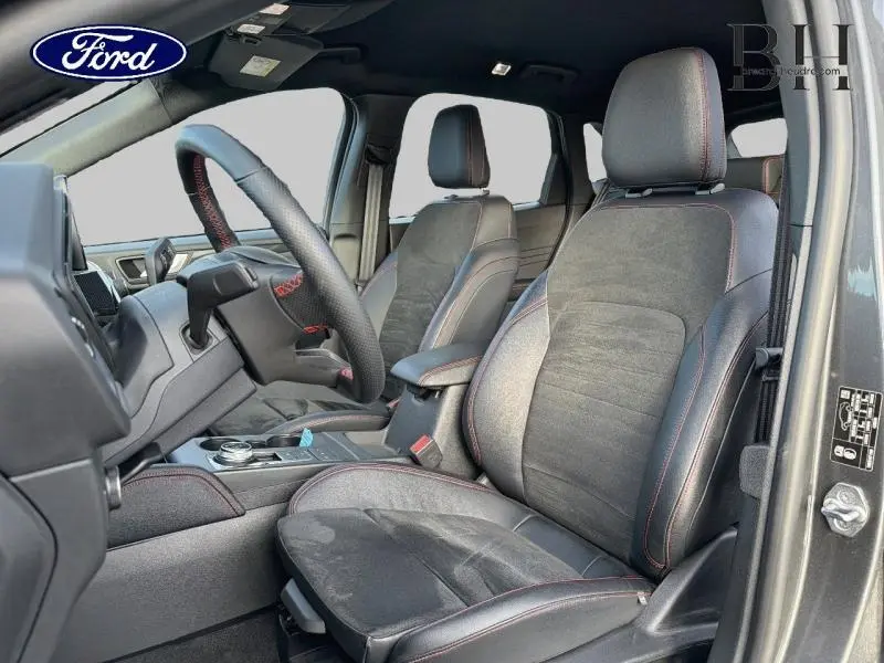 Vue intérieure côté conducteur du Ford Kuga 2023 gris Magnetic, sièges sport noirs avec surpiqûres rouges et volant cuir multifonction.