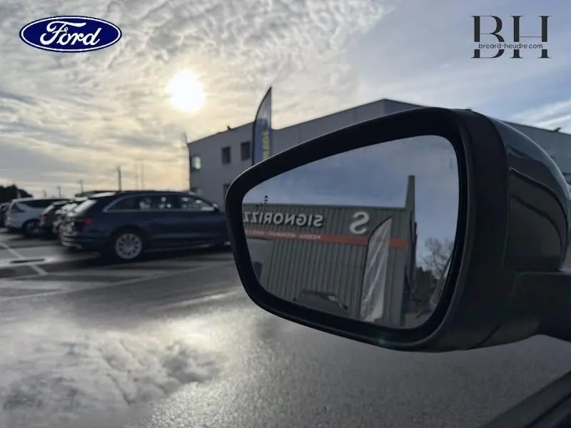 Rétroviseur côté droit d'un Ford Kuga gris Magnetic Métallisé, avec reflet d'un bâtiment et ciel nuageux en arrière-plan.