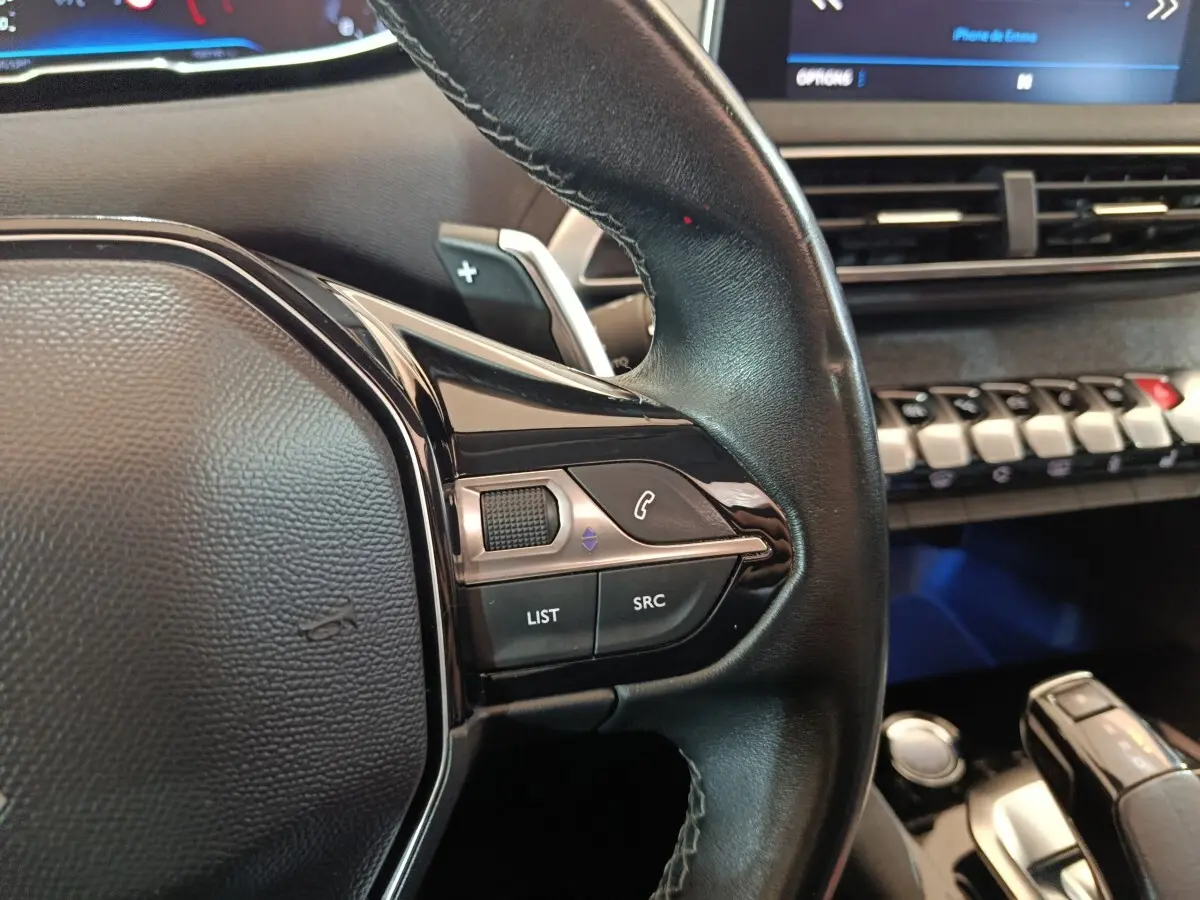 Gros plan sur le volant cuir côté gauche du Peugeot 3008 blanc, avec commandes audio et tableau de bord digital en arrière-plan.