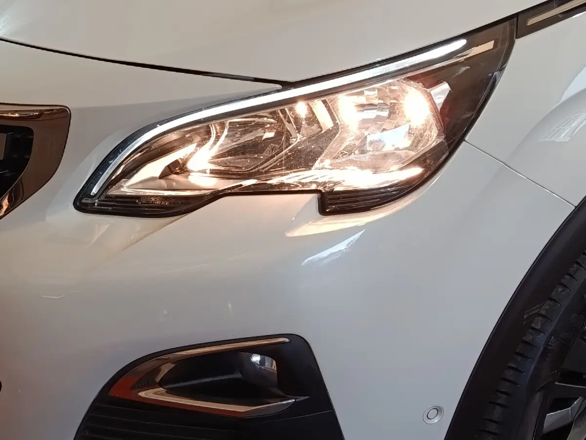 Gros plan sur le phare avant droit allumé d’un Peugeot 3008 blanc, avec antibrouillard et partie de la roue visible.