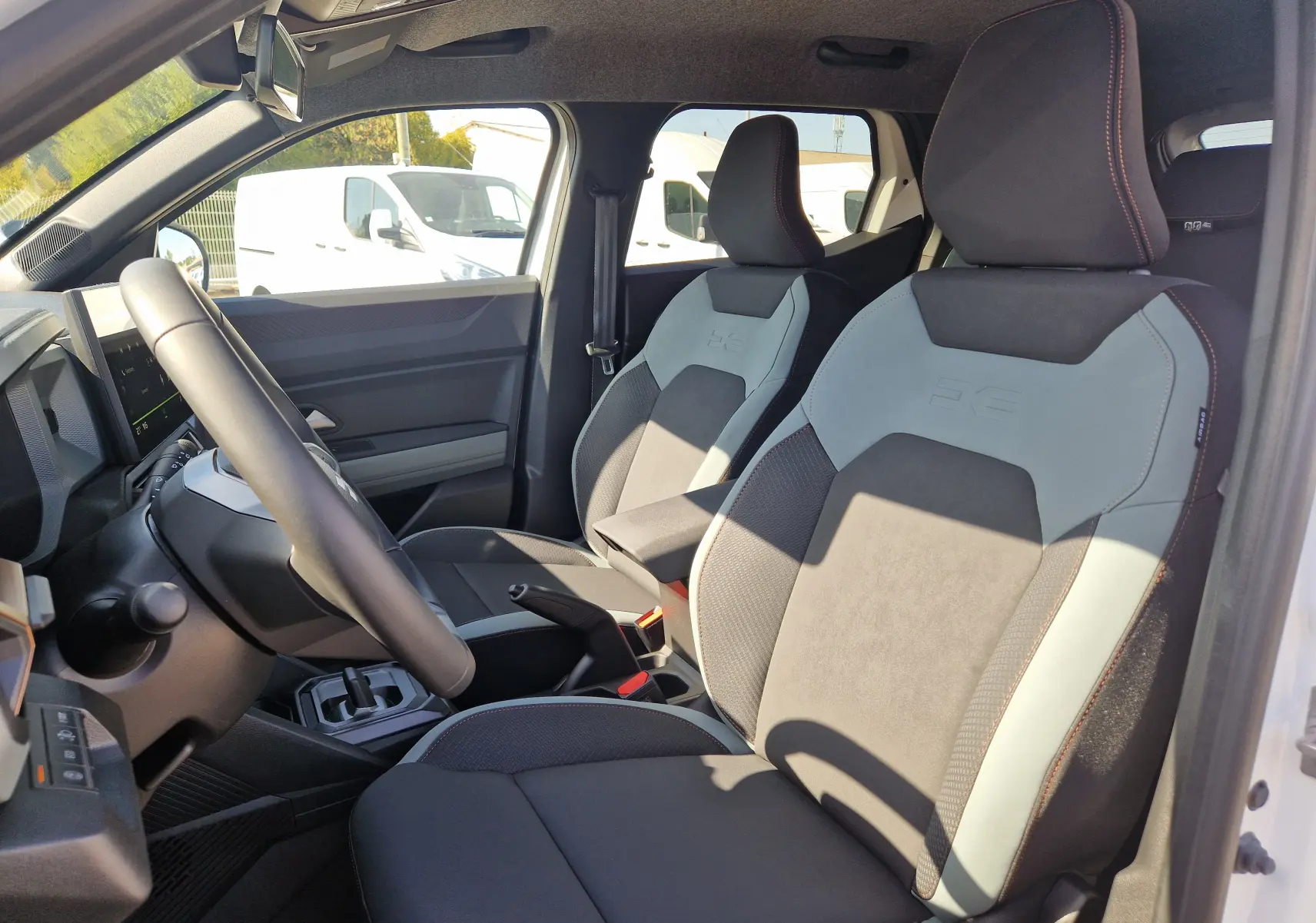 Vue intérieure côté conducteur du Dacia Duster blanc 2026, mettant en valeur les sièges bi-ton gris et noir avec surpiqûres rouges.