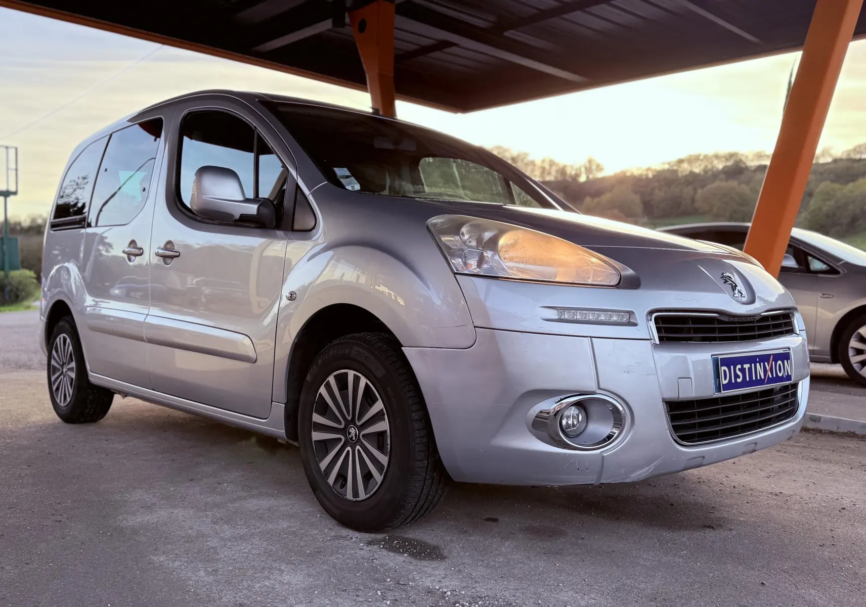 Vue 3/4 avant gauche d'un Peugeot Partner Tepee gris 2014 avec phares allumés sous un abri en fin de journée.