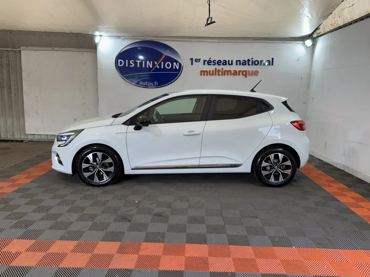 Profil droit d'une Renault Clio blanche 2023 Business TCe 100 GPL, avec jantes alliage et rétroviseurs noirs dans un garage.