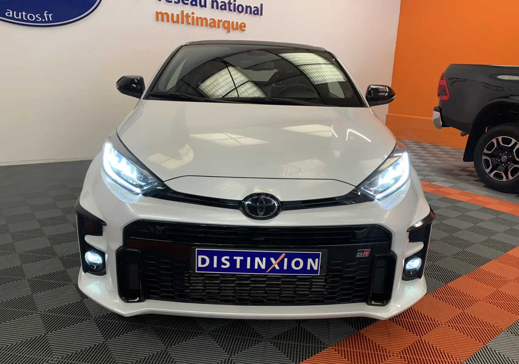 Vue avant d'une Toyota Yaris 1.6 VVT-I 261 Premium GR blanc métal avec phares LED allumés en showroom.