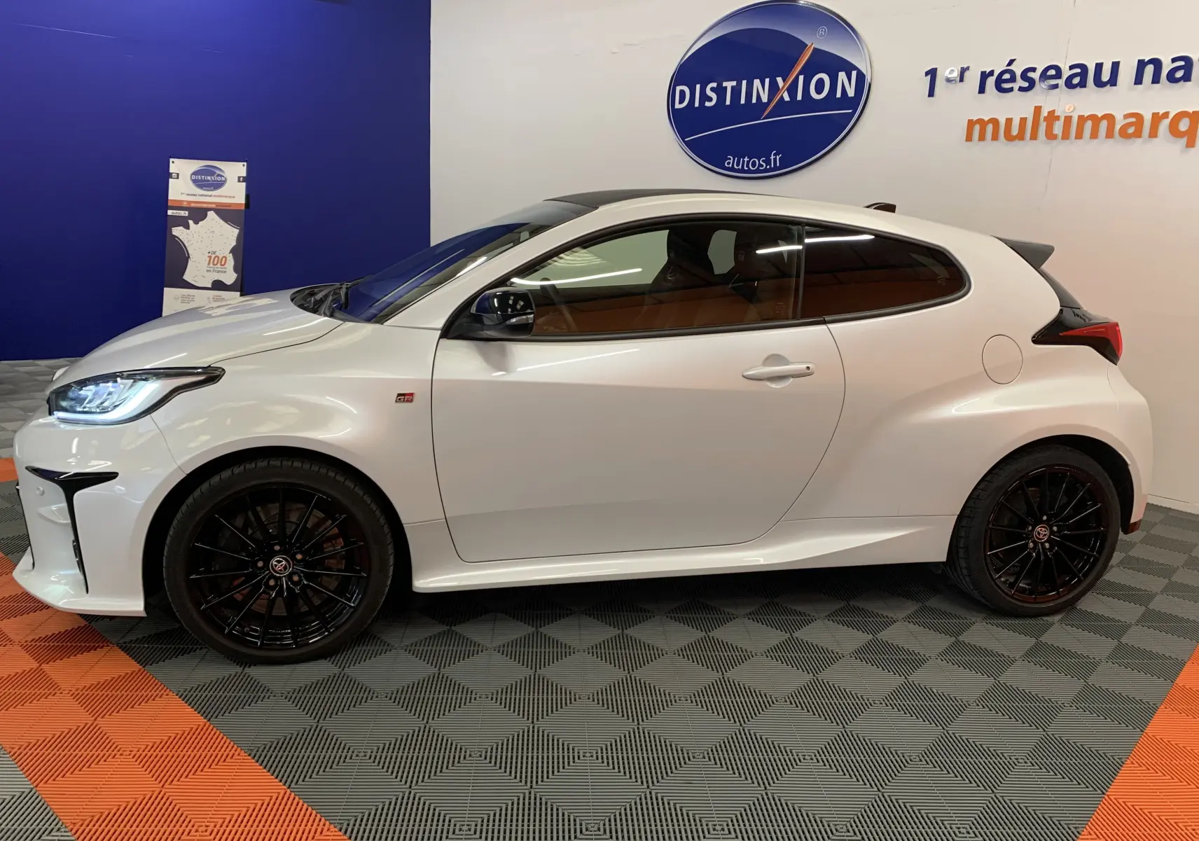 Profil côté gauche d'une Toyota Yaris 1.6 VVT-I 261 Premium GR blanche métal avec jantes noires en showroom.