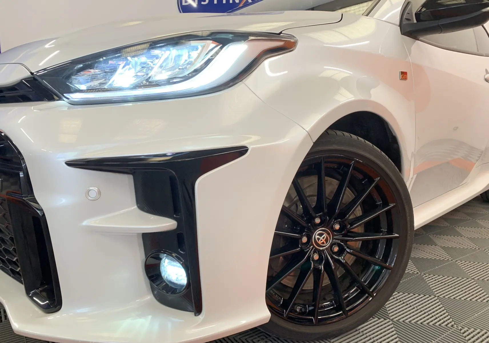 Gros plan sur l'avant droit blanc métal de la Toyota Yaris GR 1.6 VVT-I avec phares LED allumés et jantes noires sport.