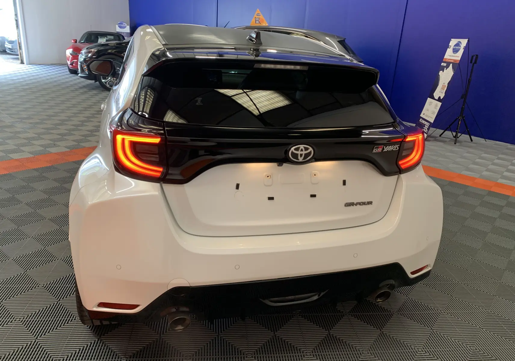 Vue arrière d'une Toyota Yaris GR blanche métal avec feux arrière allumés et toit noir dans un showroom.