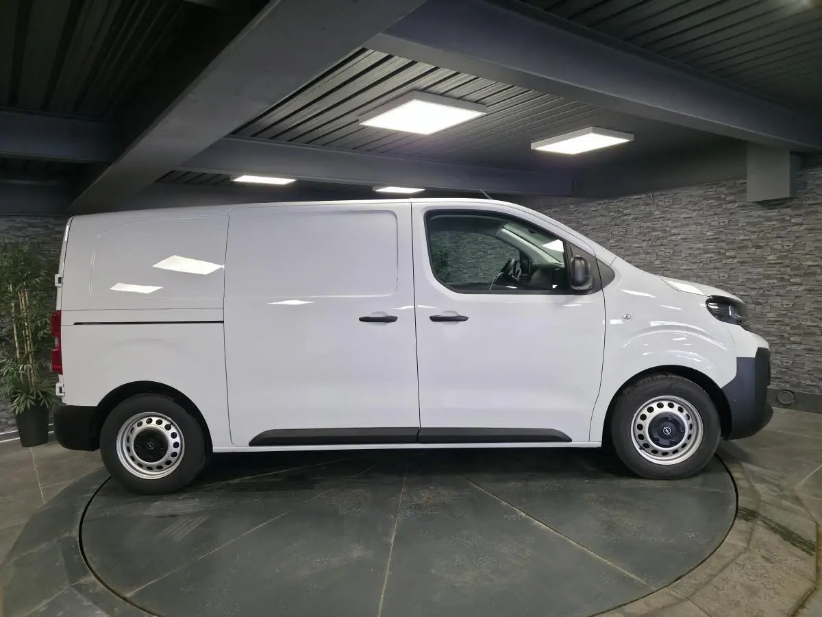 Vue de profil côté gauche d’un utilitaire Opel Vivaro blanc avec roues tôles grises en intérieur showroom.