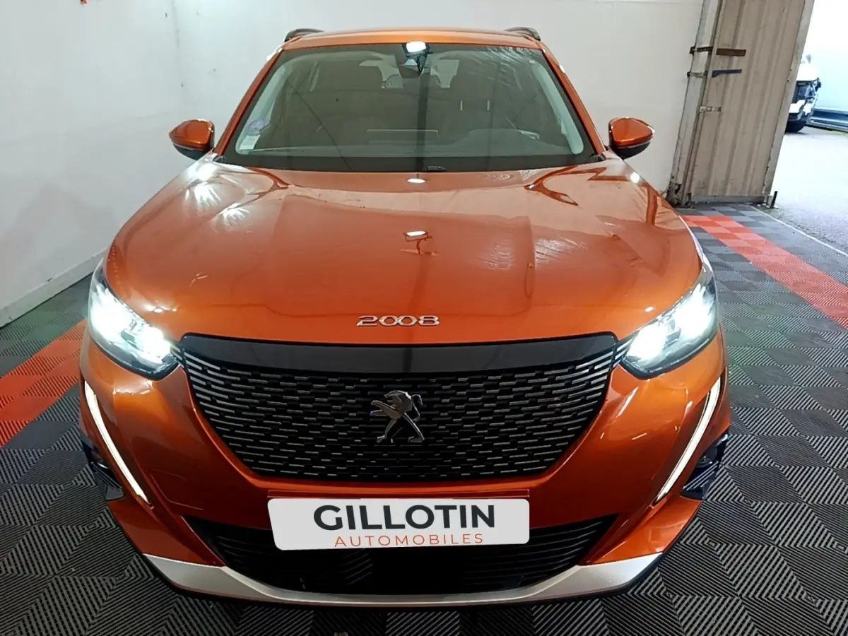 Vue avant d'un Peugeot 2008 orange avec phares LED allumés et calandre noire distinctive dans un garage.