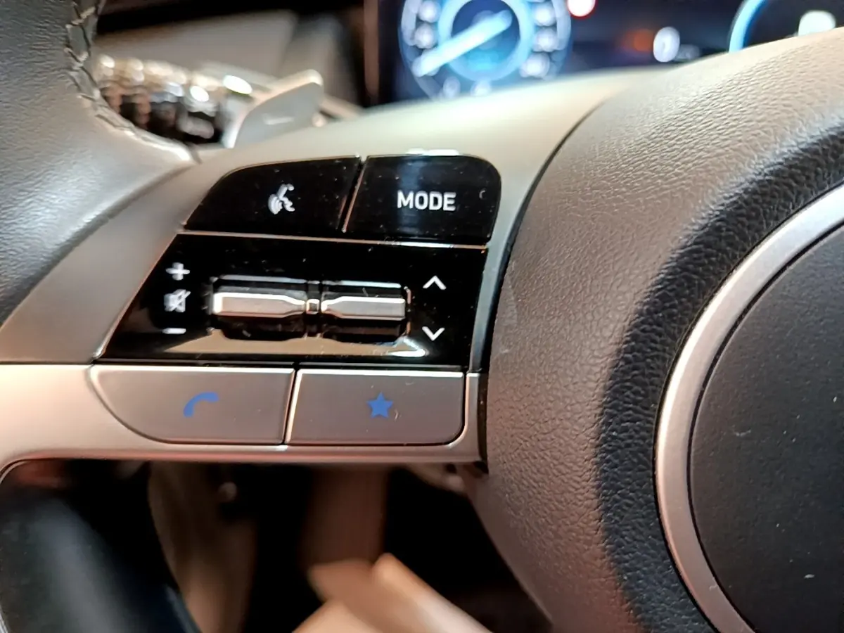 Gros plan sur les commandes gauche du volant cuir et alu du Hyundai Tucson bleu 2023, avec affichage digital en arrière-plan.