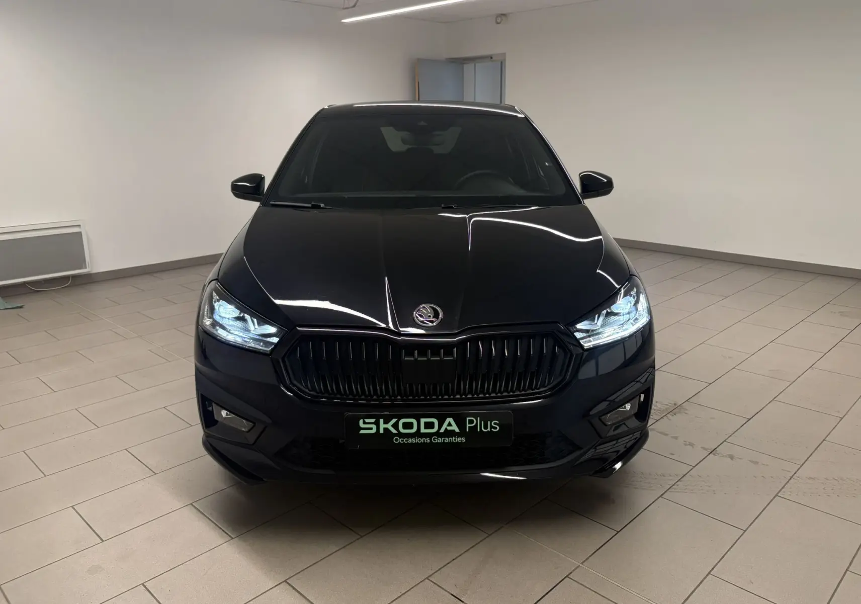 Vue avant d'une Skoda Fabia 2025 noire Magic nacré avec calandre noire et phares allumés en intérieur showroom.