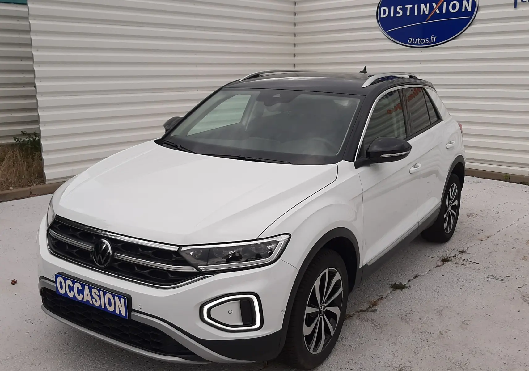 Volkswagen T-Roc blanc pur avec toit noir, vue 3/4 avant droit, stationné devant un mur blanc avec panneau Distinxion.