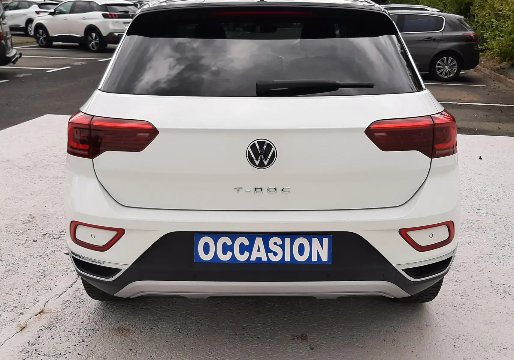 Vue arrière d'un Volkswagen T-Roc blanc pur avec toit noir, immatriculé "OCCASION", dans un parking extérieur.