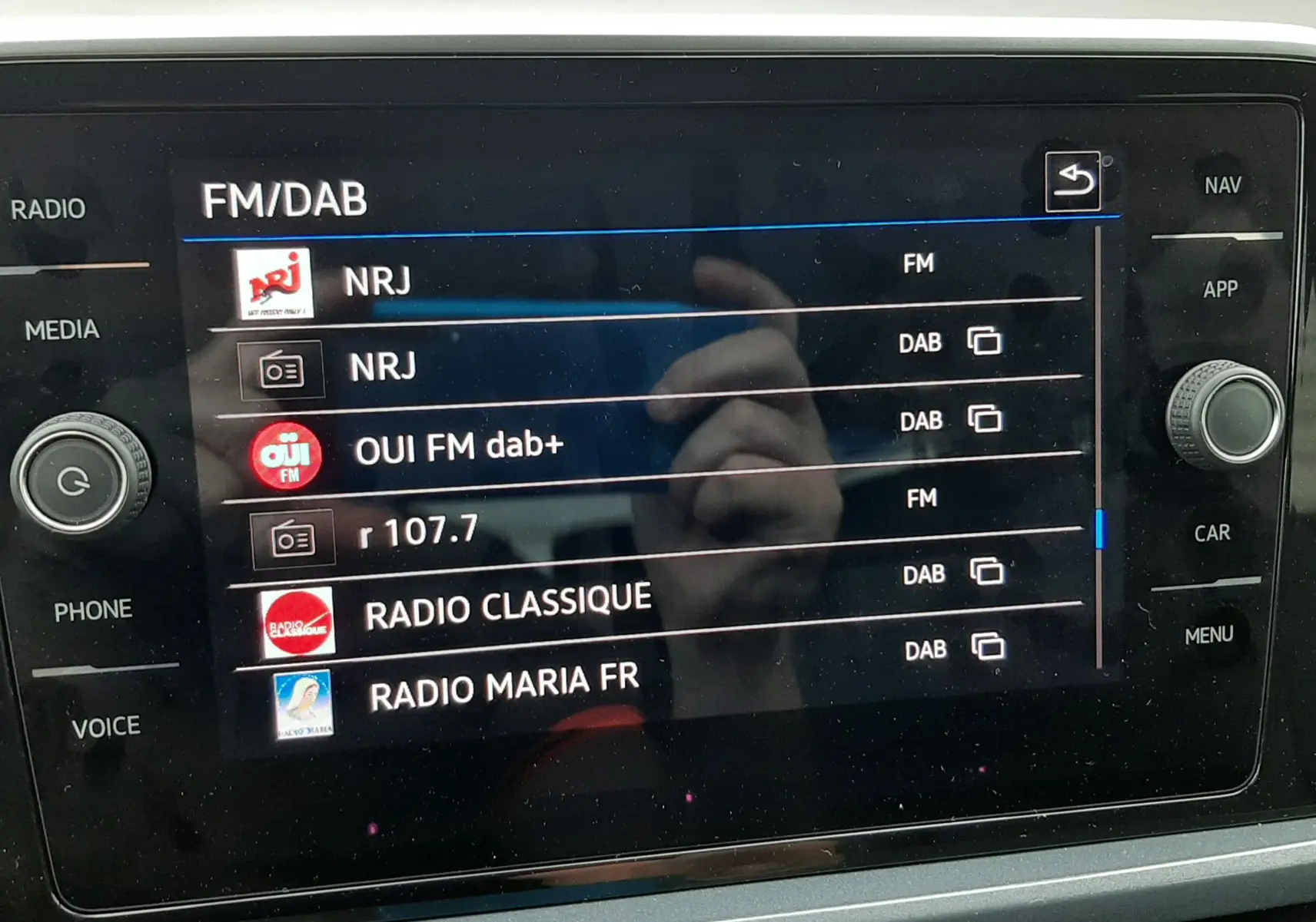 Vue rapprochée de l’écran tactile multimédia du Volkswagen T-Roc 2.0 TDI blanc avec toit noir, affichant les stations radio FM/DAB.