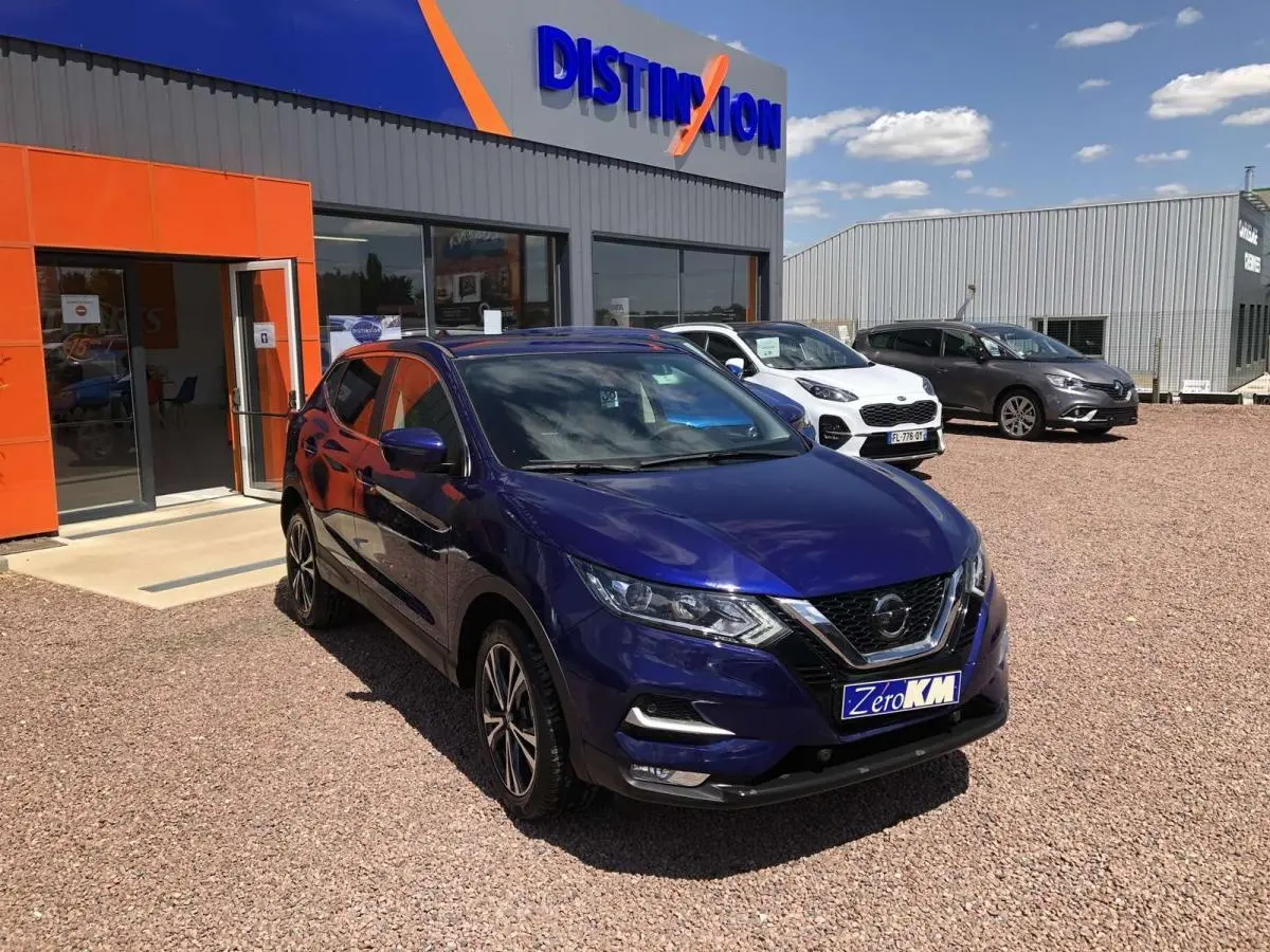 Nissan Qashqai bleu métallisé en 3/4 avant droit, stationné devant un concessionnaire avec panneau Distinxion visible.