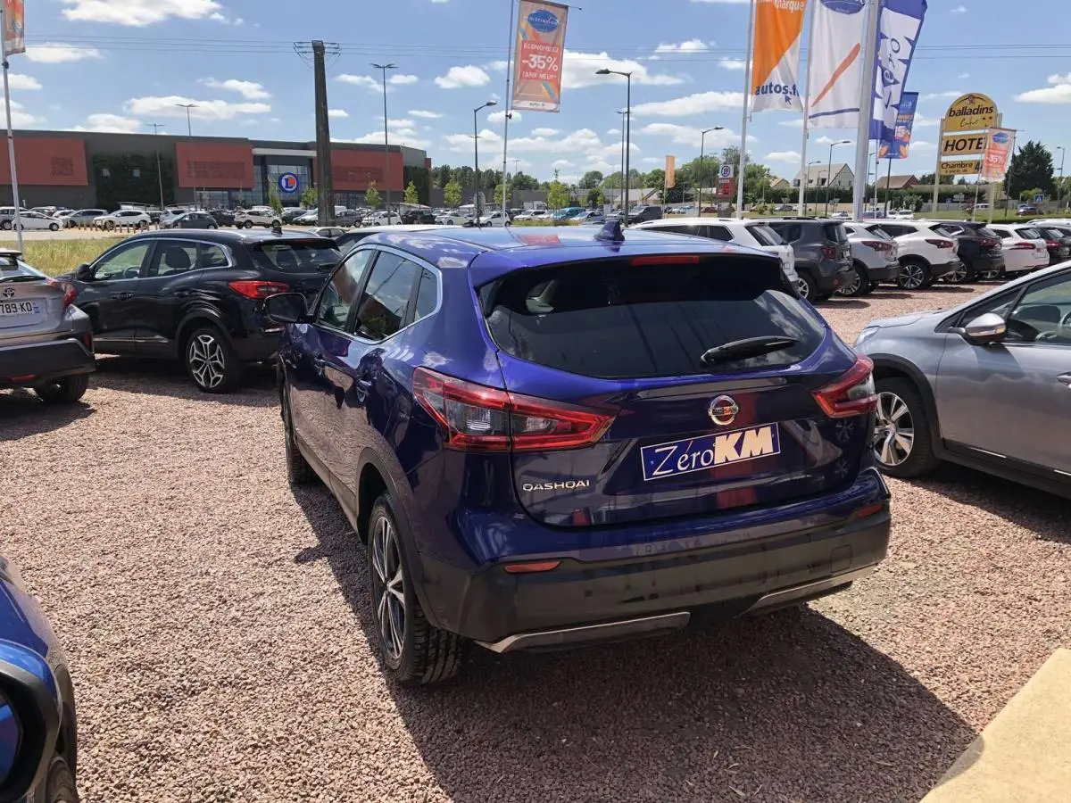 Vue 3/4 arrière d'un Nissan Qashqai bleu métallisé garé sur un parking extérieur sous un ciel ensoleillé.