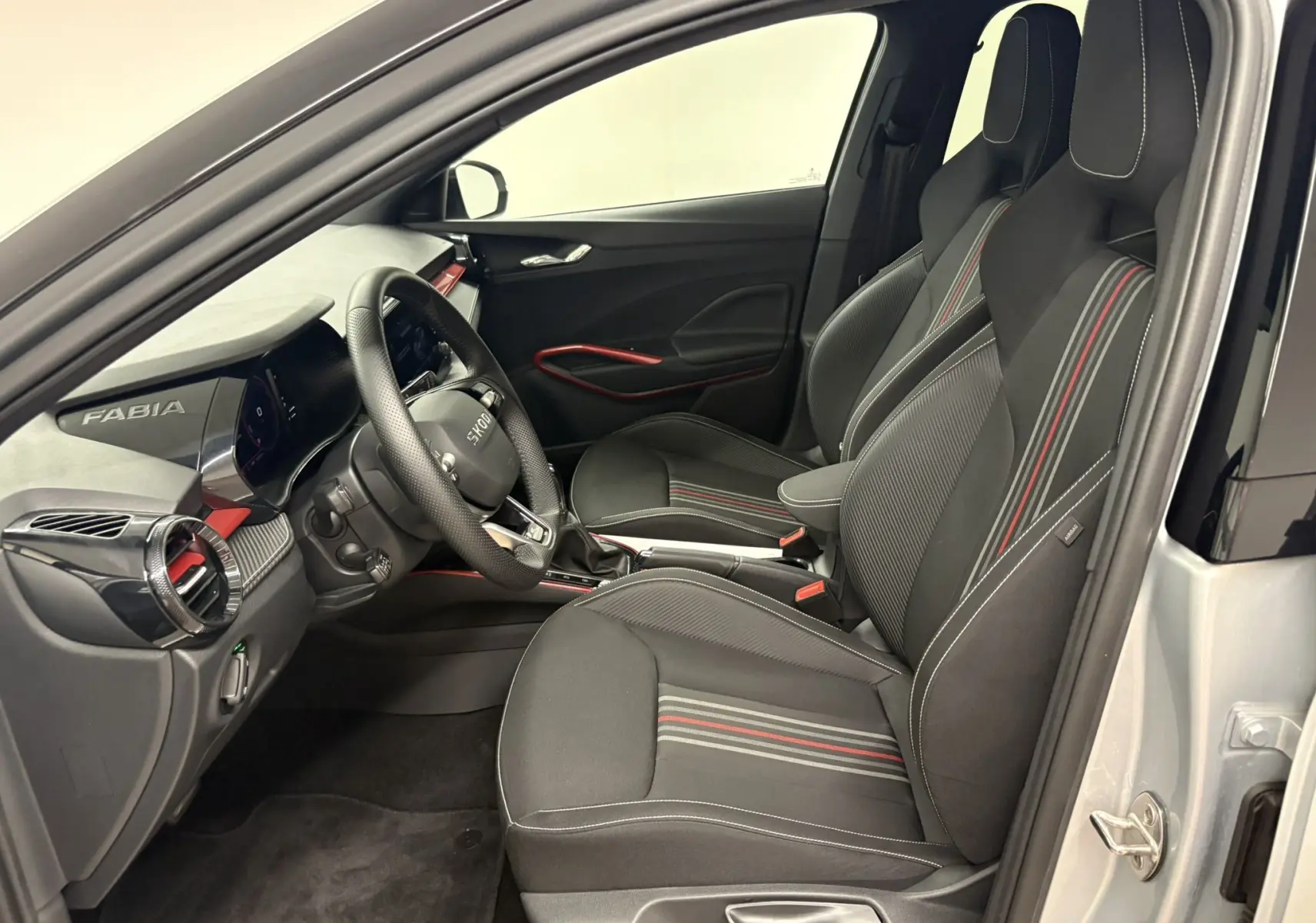 Vue intérieure côté conducteur de la Skoda Fabia 2025 gris argent avec sièges sport noirs à bandes rouges et tableau de bord moderne.