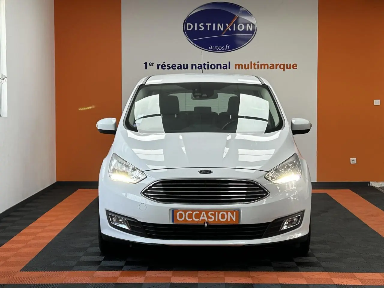 Vue frontale d'une Ford C-MAX blanche 2018 avec calandre chromée et phares allumés dans un showroom.