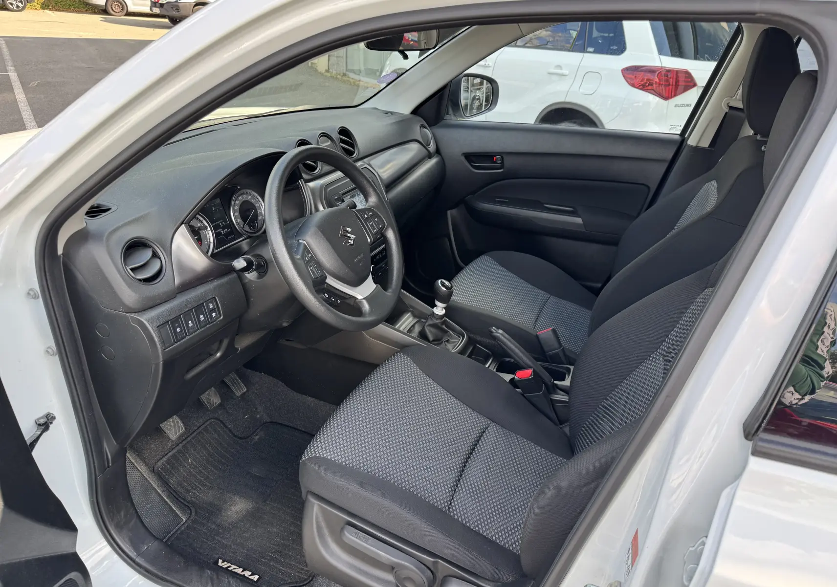 Vue intérieure côté conducteur du Suzuki Vitara blanc 2023, montrant le tableau de bord et les sièges en tissu gris et noir.