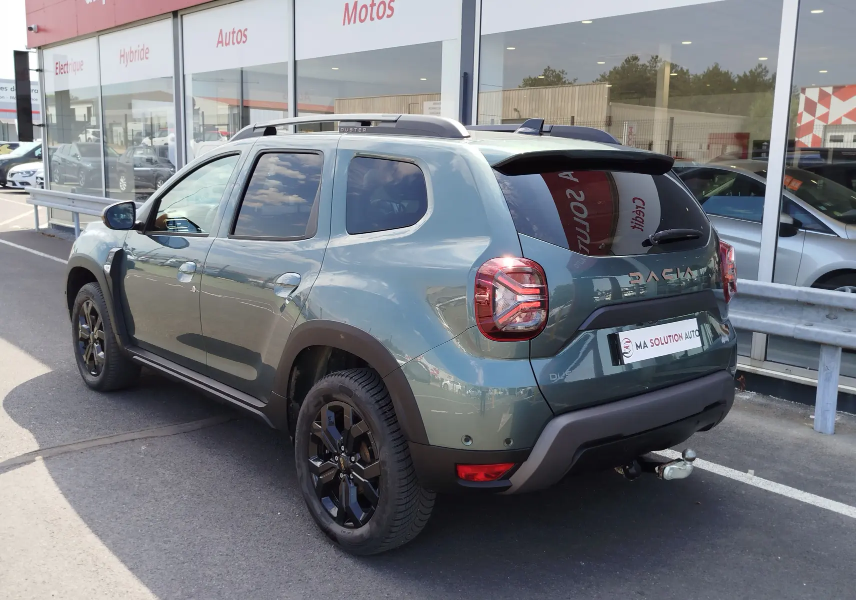 Vue 3/4 arrière droite du Dacia Duster 2024 Vert Cèdre métallisé avec jantes noires et attelage visible.