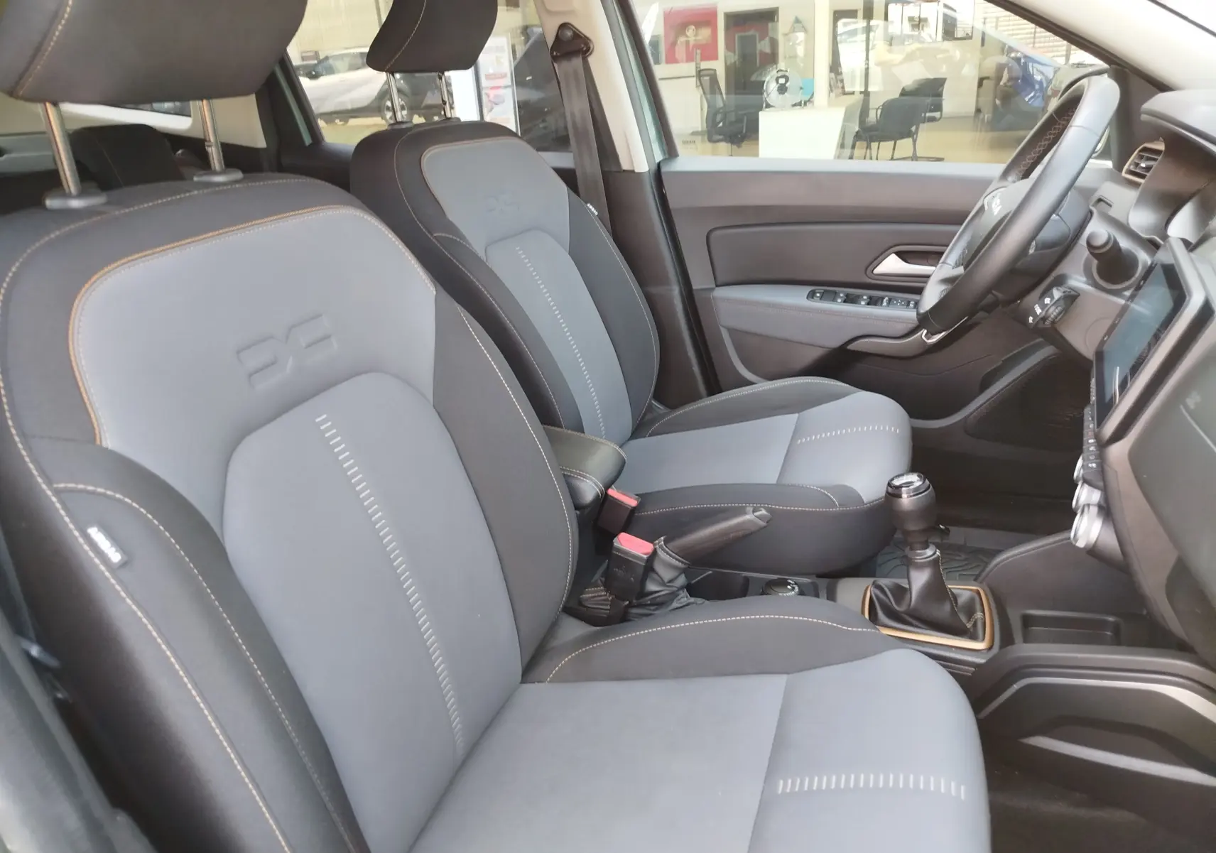 Vue intérieure côté conducteur du Dacia Duster 2024, sièges gris avec surpiqûres orange et levier de vitesse manuel visible.