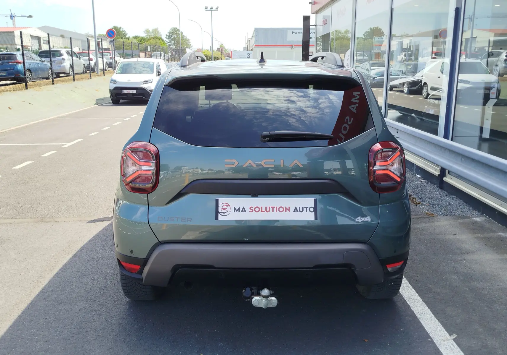 Vue arrière d’un Dacia Duster 2024 Vert Cèdre métallisé avec logo DACIA et attelage visible sur un parking extérieur.