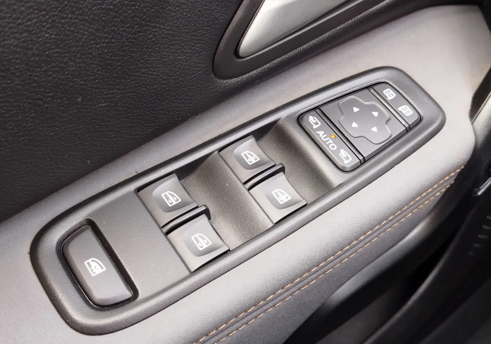 Gros plan sur les commandes des vitres électriques et des rétroviseurs côté conducteur d'un Dacia Duster 2024, garniture noire avec surpiqûres marron.