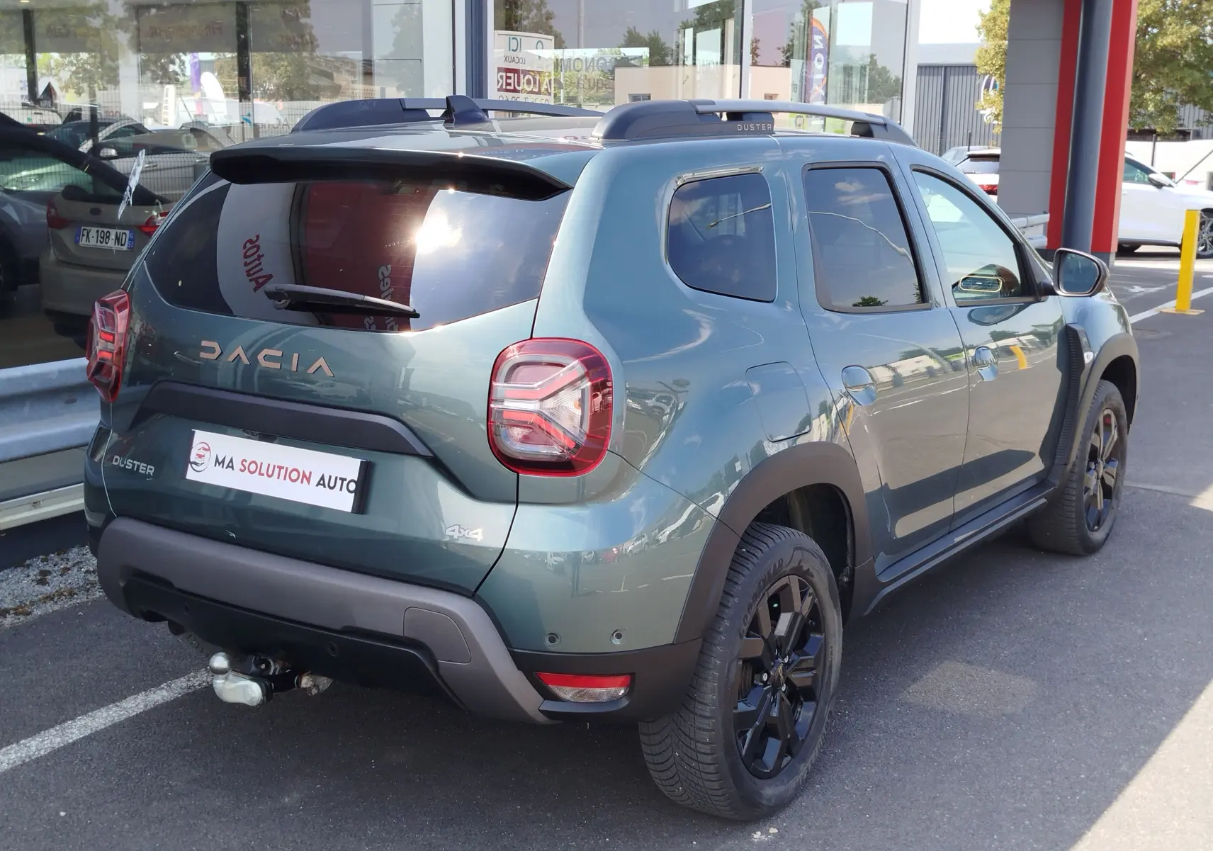 Vue 3/4 arrière droite d'un Dacia Duster 2024 Vert Cèdre métallisé avec jantes noires et attelage visible.