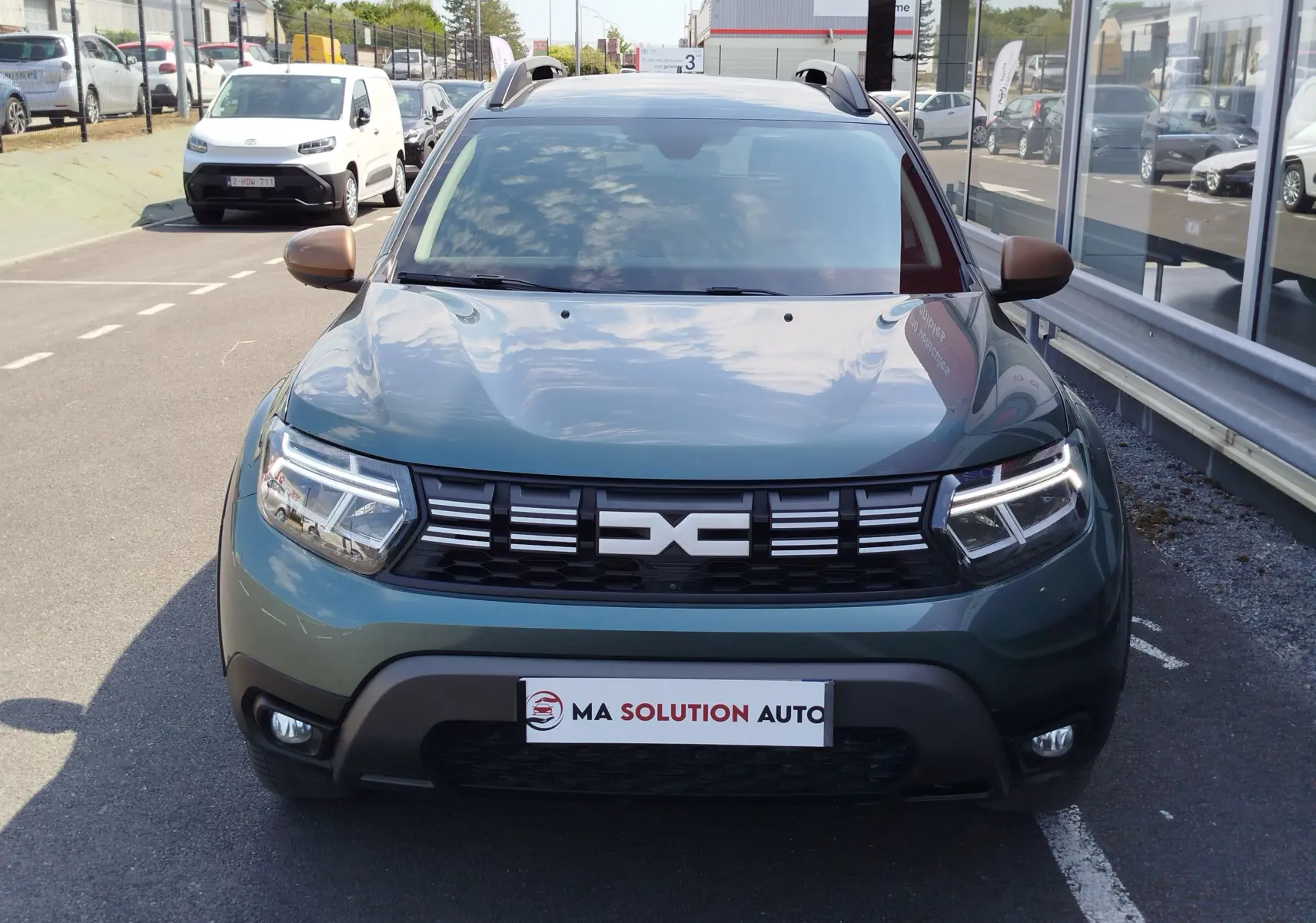 Vue de face d'un Dacia Duster 2024 vert cèdre métallisé avec calandre noire et feux LED allumés.
