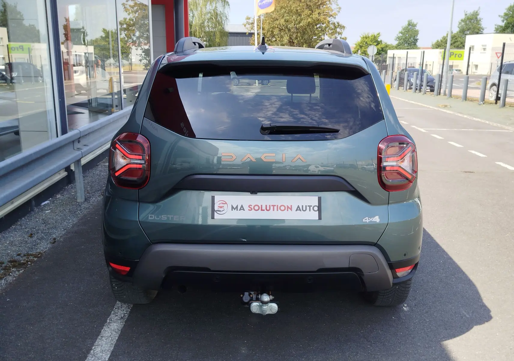 Vue arrière d'un Dacia Duster 2024 Vert Cèdre métallisé avec logo DACIA et attelage visible en stationnement.