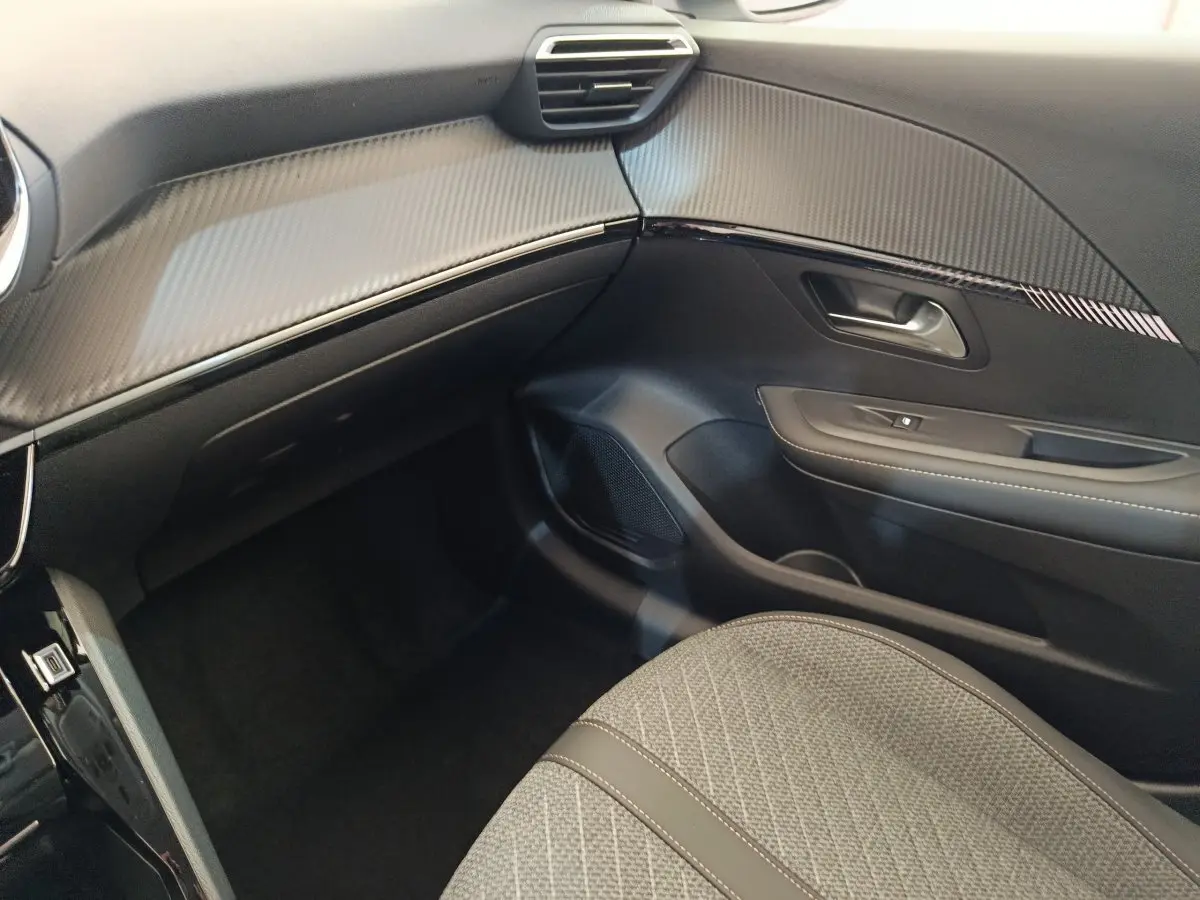 Vue intérieure côté passager de la Peugeot 208 électrique 2024 avec tableau de bord noir et siège tissu gris.