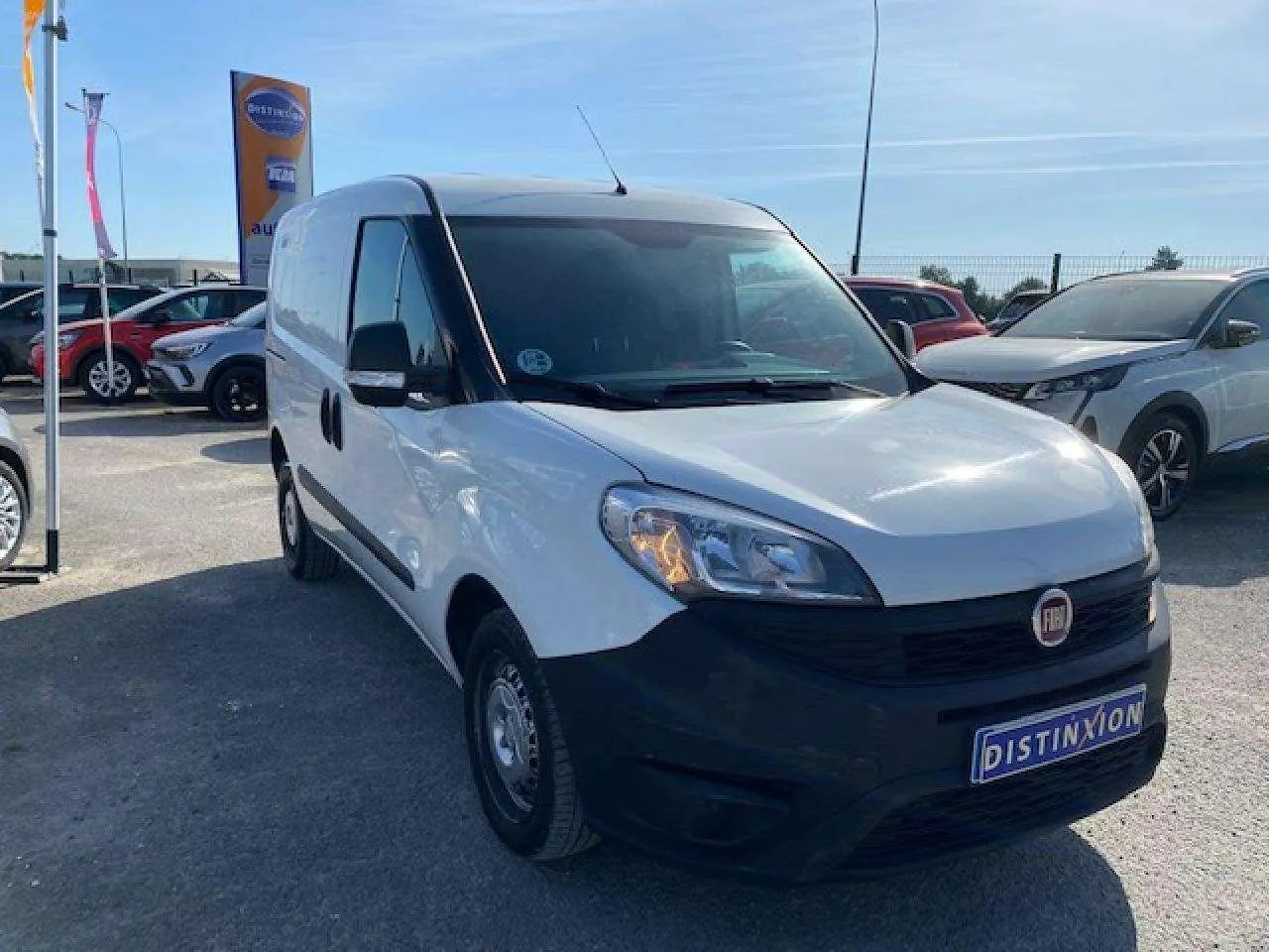 Fiat Doblo blanc vu en 3/4 avant droit, fourgon tôlé avec pare-chocs noirs et porte latérale droite visible.