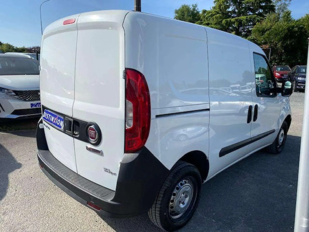 Fiat Doblo blanc vu en 3/4 arrière droit, fourgon tôlé avec pare-chocs noirs et porte latérale droite visible.