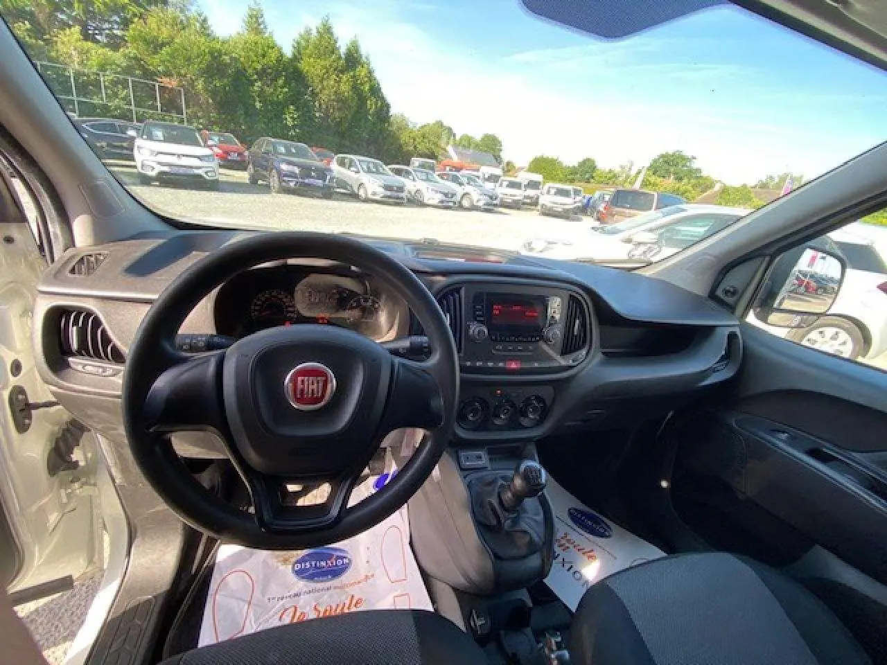 Vue intérieure du poste de conduite du FIAT Doblo blanc de 2017, avec volant, tableau de bord et levier de vitesses manuelle visibles.