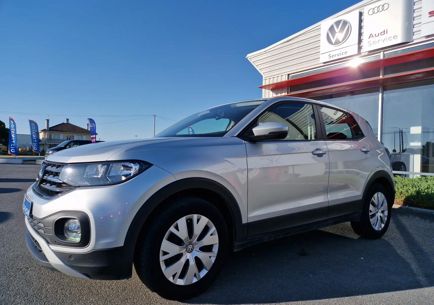 Volkswagen T-Cross argent métal vu de 3/4 avant droit, stationné devant un service Volkswagen et Audi par temps clair.