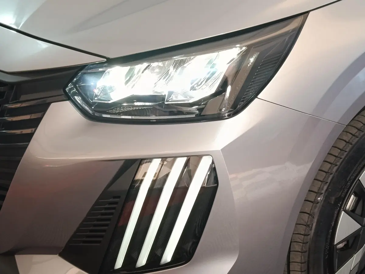 Gros plan sur l'avant droit gris clair du Peugeot 208 électrique 2024, mettant en valeur ses phares LED et feux diurnes verticaux.