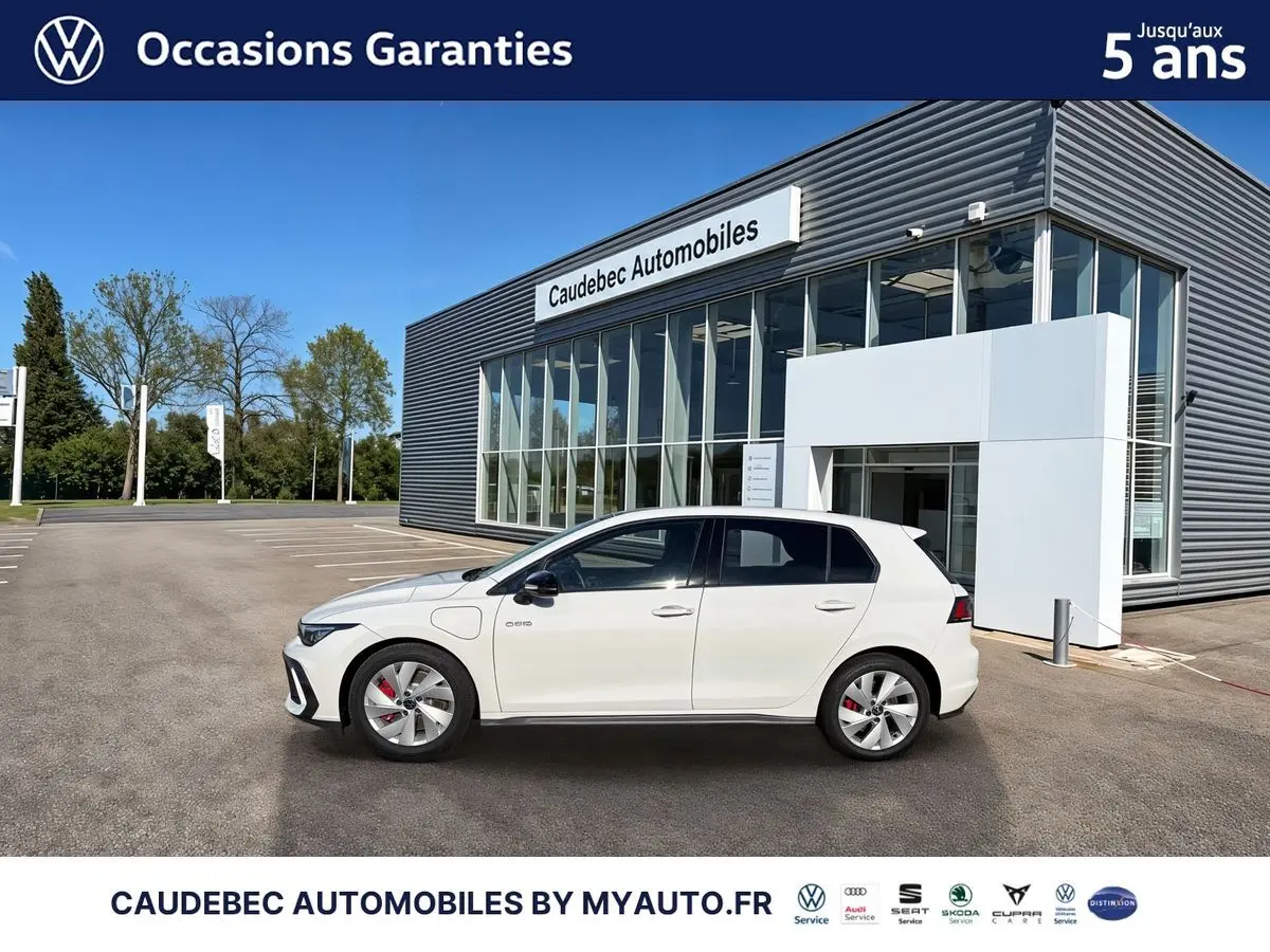 Vue de profil côté gauche d'une Volkswagen Golf 1.5 eHybrid 272 DSG6 blanche stationnée devant un bâtiment moderne.