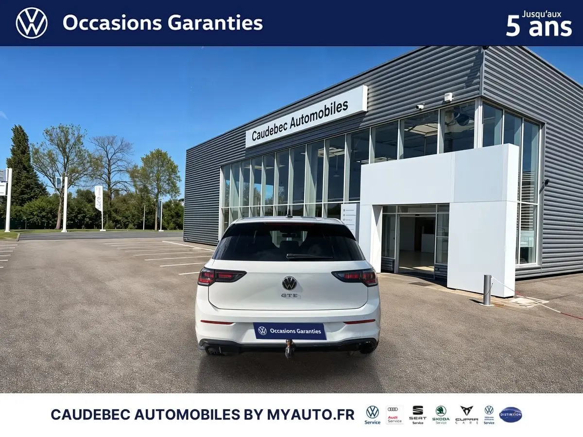 Vue arrière d'une Volkswagen Golf 1.5 eHybrid blanche avec attelage escamotable, stationnée devant un concessionnaire.