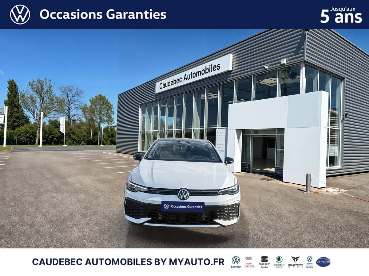 Vue frontale d'une Volkswagen Golf 1.5 eHybrid 272 DSG6 blanche, devant un bâtiment moderne de concession automobile.