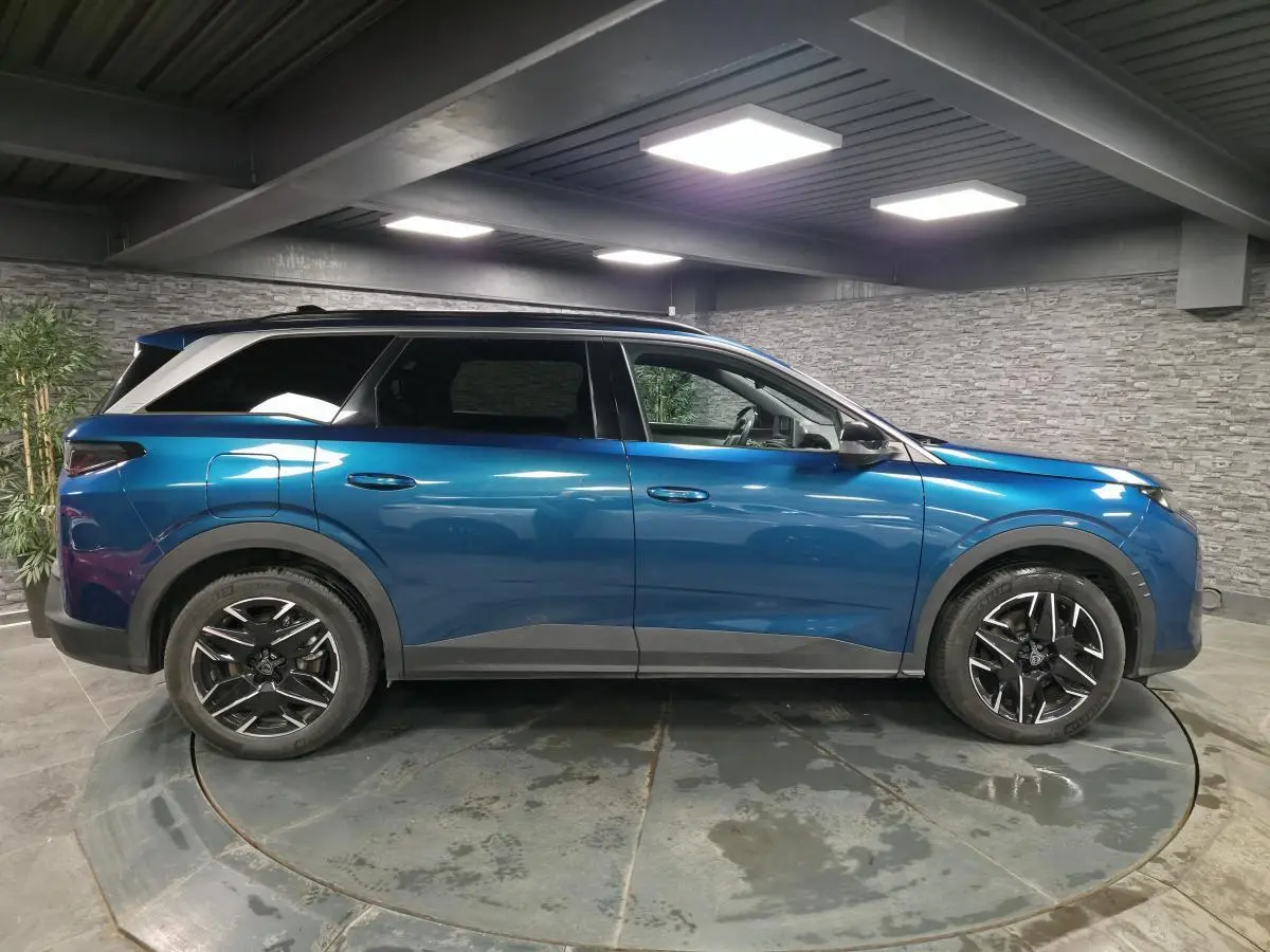 Profil droit d’un Peugeot 5008 bleu métallisé 2025 en intérieur avec jantes alliage noires et toit noir.
