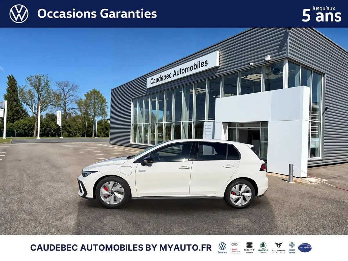 Vue latérale droite d'une Volkswagen Golf 1.5 eHybrid blanche garée devant un bâtiment moderne en verre.